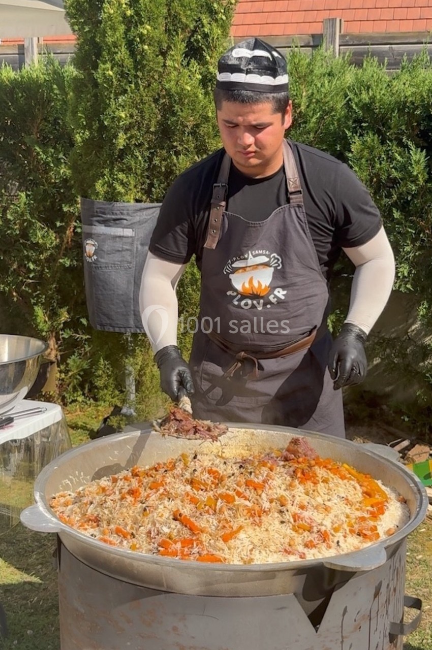 Un cuisinier prépare un grand plat de riz avec des légumes et de la viande dans un récipient métallique en extérieur.