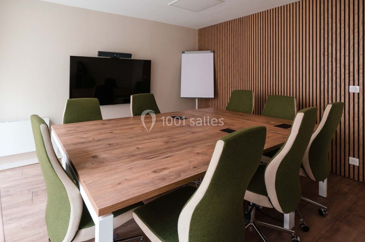 Salle de réunion avec table en bois, chaises vertes, écran mural, tableau blanc et mur en bois à l'arrière.