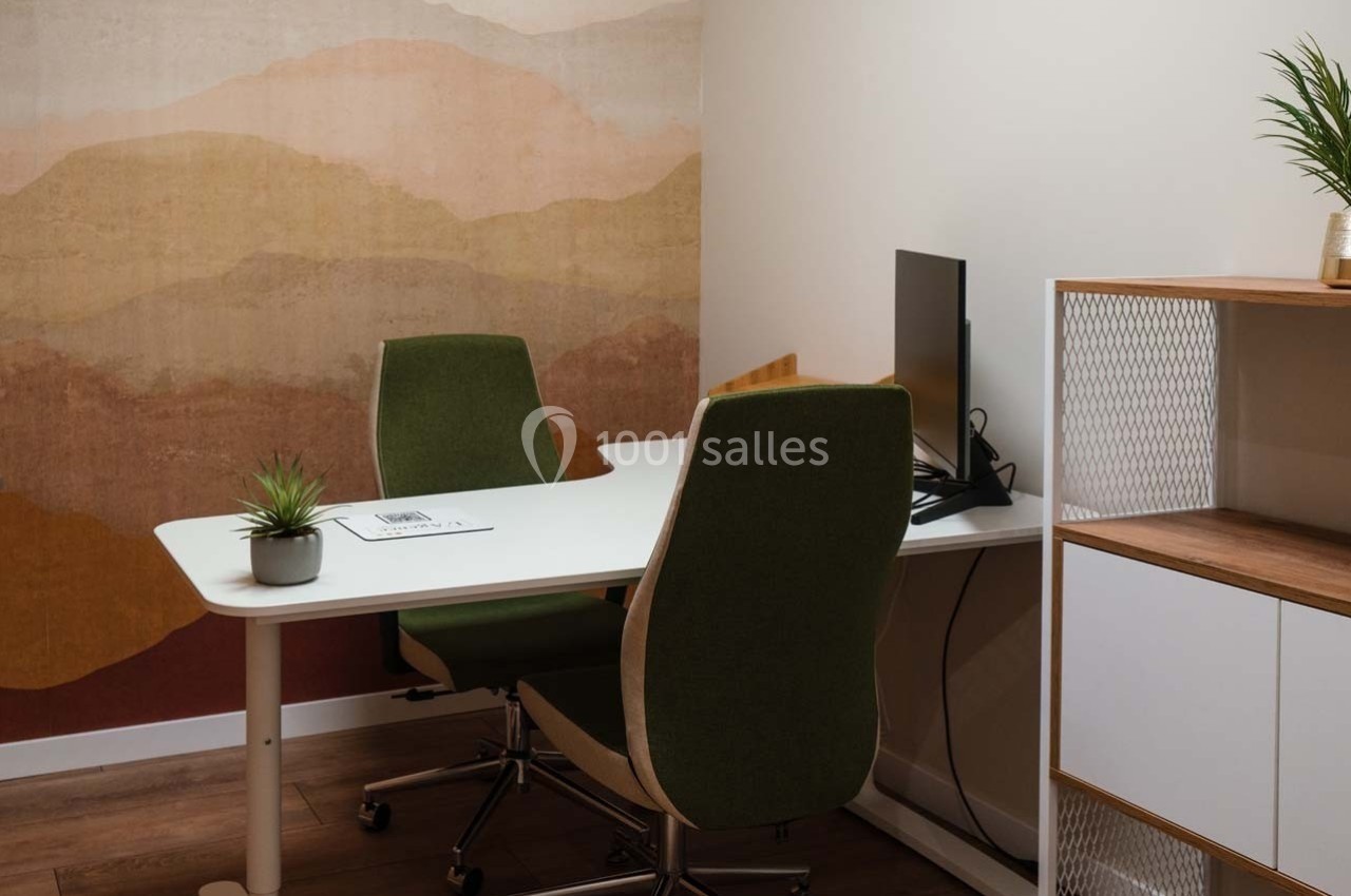 Bureau avec deux chaises vertes, un écran d'ordinateur, une plante et une étagère, dans une pièce au mur décoratif.