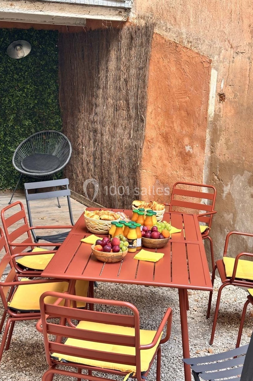 Table extérieure rouge entourée de chaises jaunes, garnie de fruits, jus et pain, dans une cour avec murs en pierre et…
