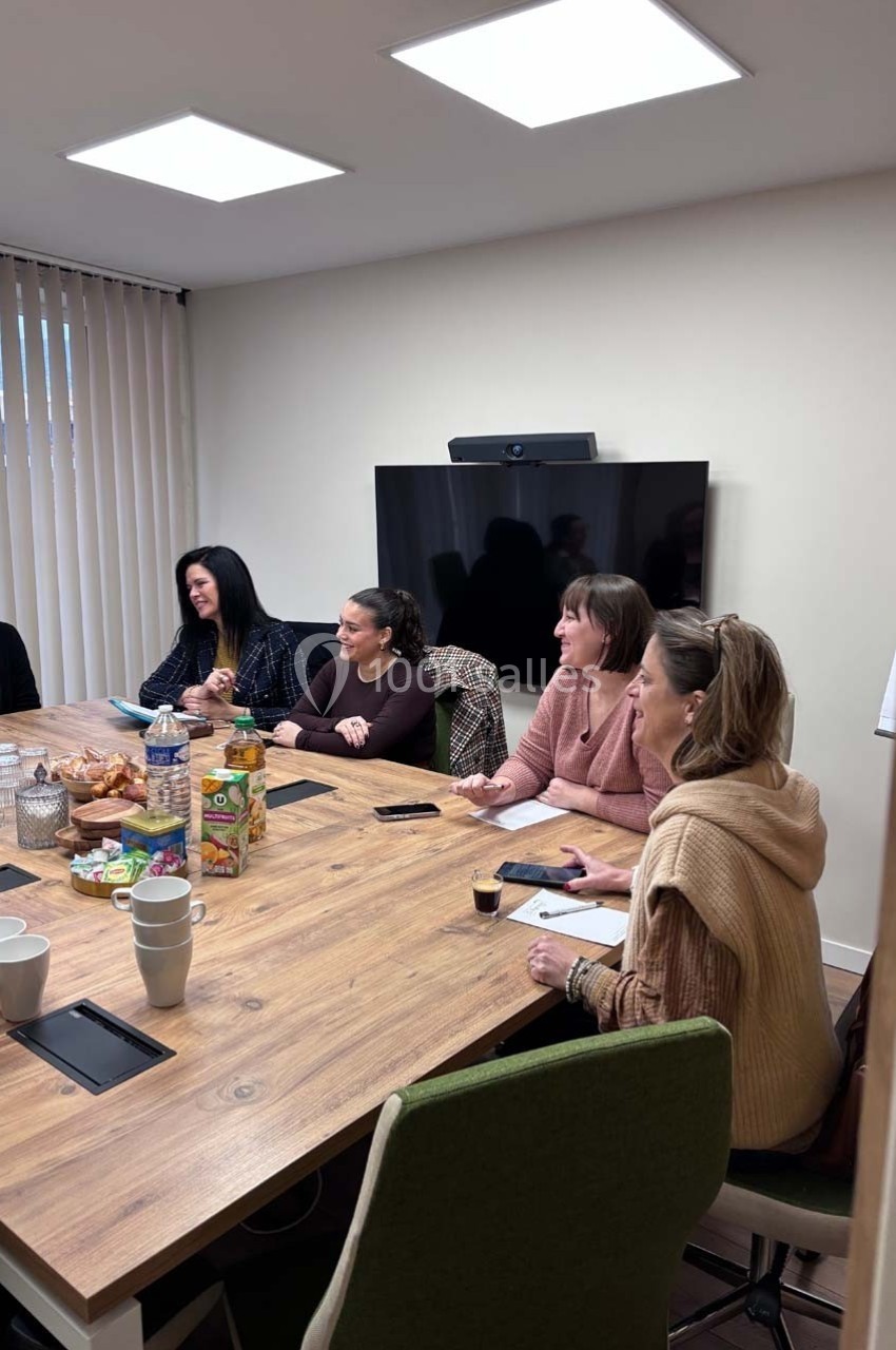 Des personnes assises autour d'une table en bois lors d'une réunion dans une salle équipée d'un écran.
