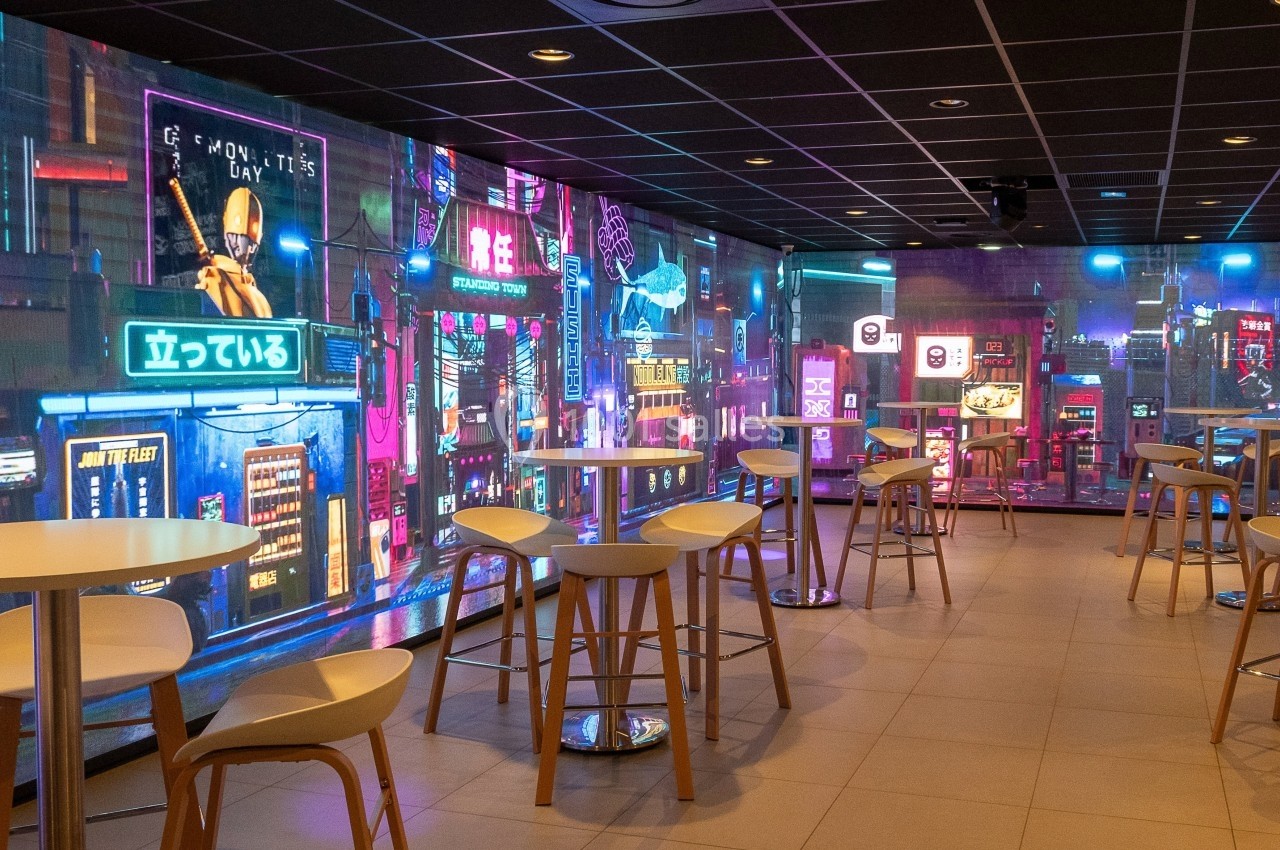 Salle moderne avec tables hautes et tabourets, décorée de murs lumineux affichant des néons et paysages urbains futuristes.
