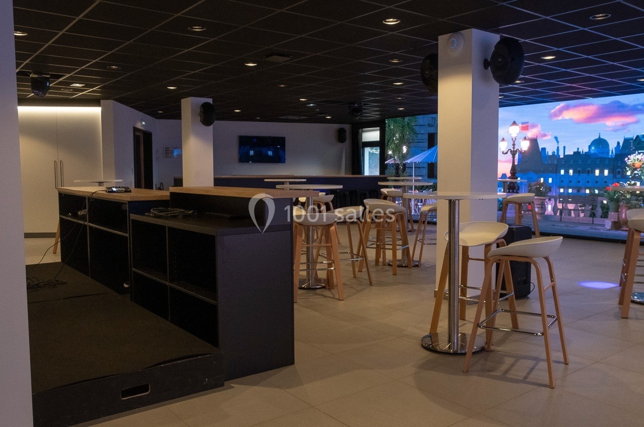 Location salle Marines (Val-d'Oise) - Stand Ing Events #21 Salle moderne avec tables hautes, tabourets et grande baie vitrée donnant sur une vue urbaine éclairée.