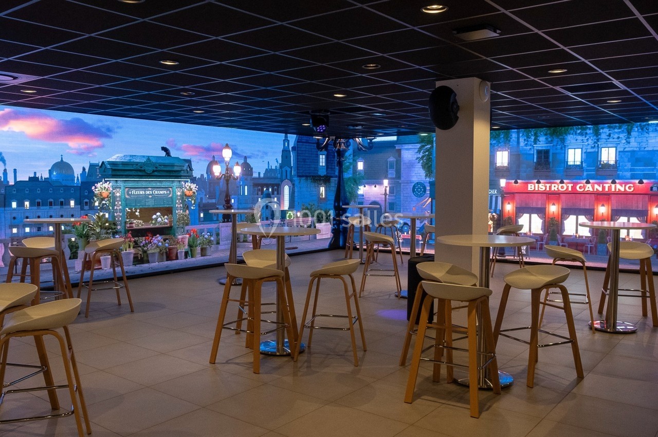 Location salle Marines (Val-d'Oise) - Stand Ing Events #28 Terrasse intérieure avec tables et tabourets, vue sur une façade lumineuse de restaurant et un décor urbain.