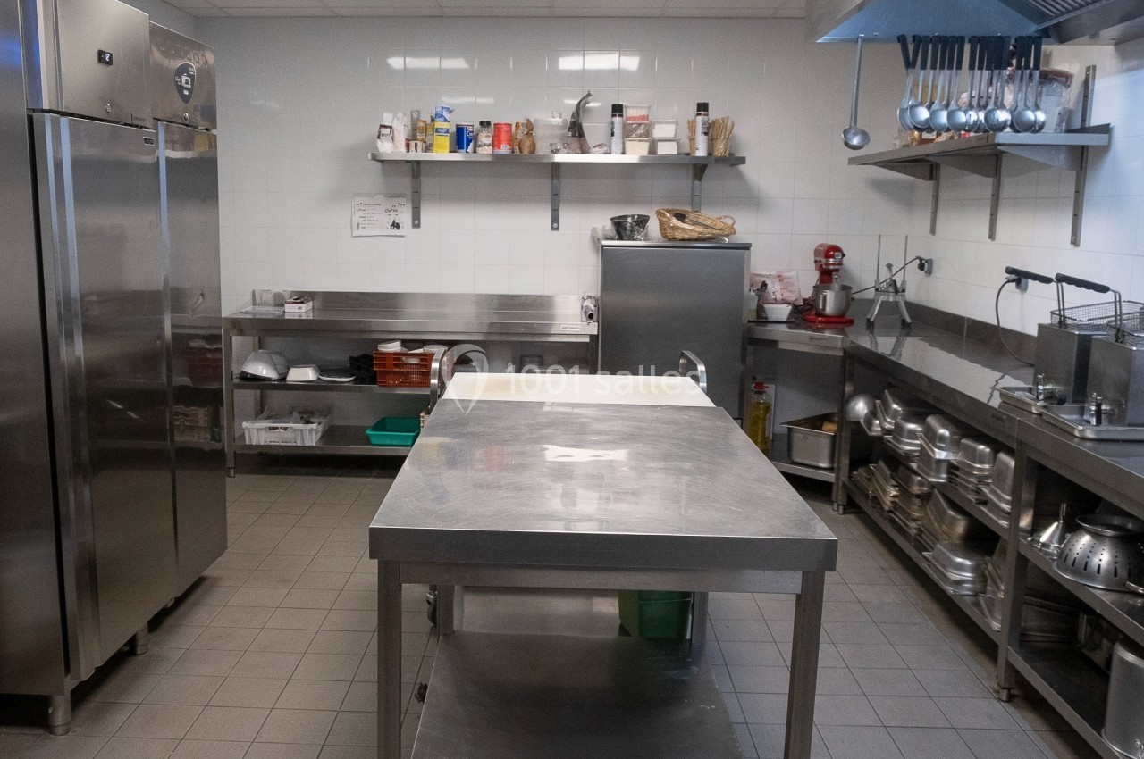Cuisine professionnelle avec plan de travail en inox, étagères de rangement, réfrigérateur et ustensiles suspendus.