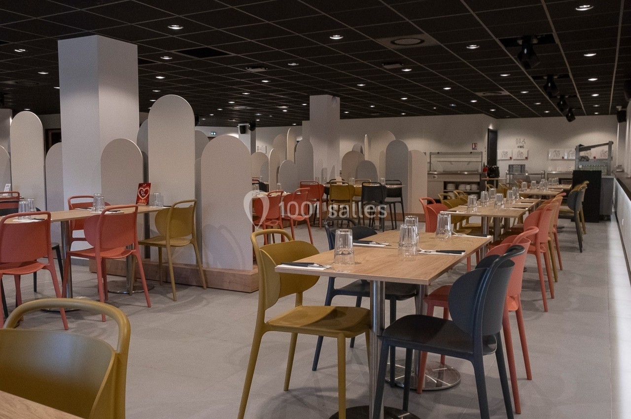 Salle de restaurant moderne avec tables en bois, chaises colorées et séparateurs blancs sous un plafond sombre.