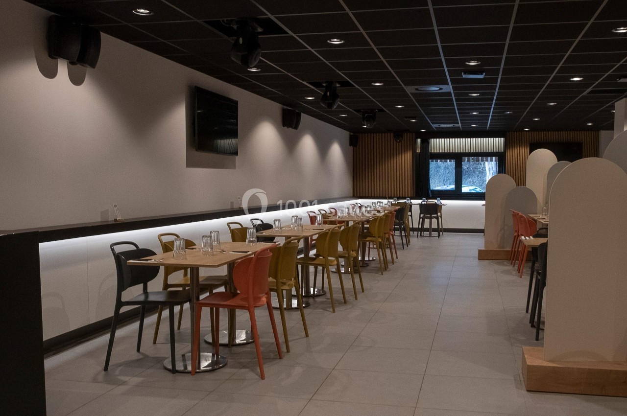 Salle de restaurant moderne avec des tables alignées, chaises colorées et éclairage tamisé.
