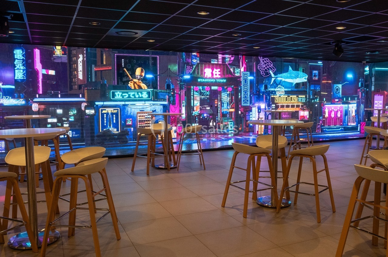 Salle moderne avec tables et tabourets hauts, devant un mur d'écrans affichant une ville futuriste éclairée par des néons.