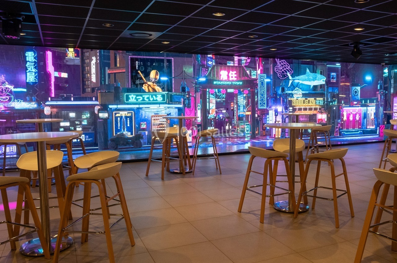 Salle moderne avec tables hautes et tabourets, décorée d'un écran mural affichant une rue animée de style néon futuriste.