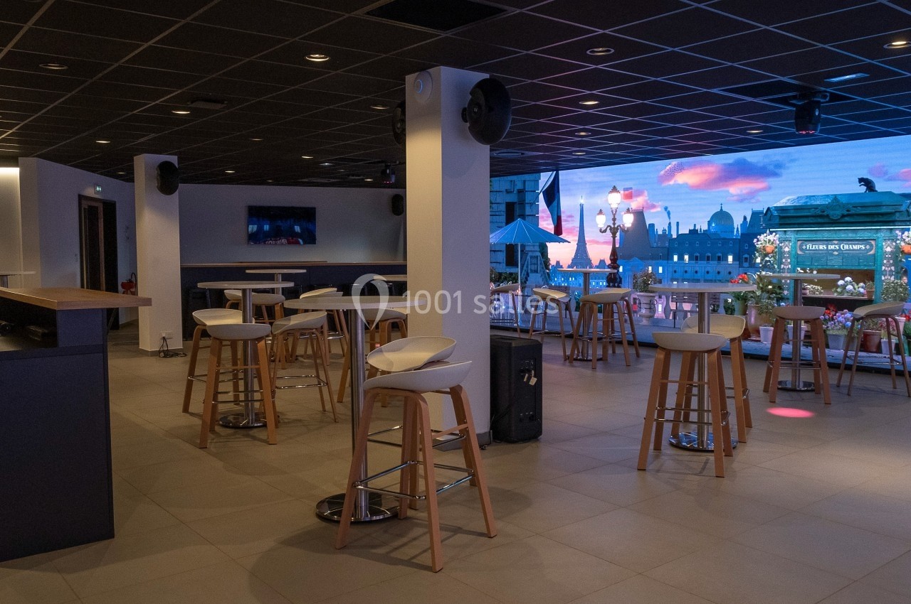 Salle moderne avec tables hautes, tabourets et écran mural affichant une vue urbaine illuminée au crépuscule.