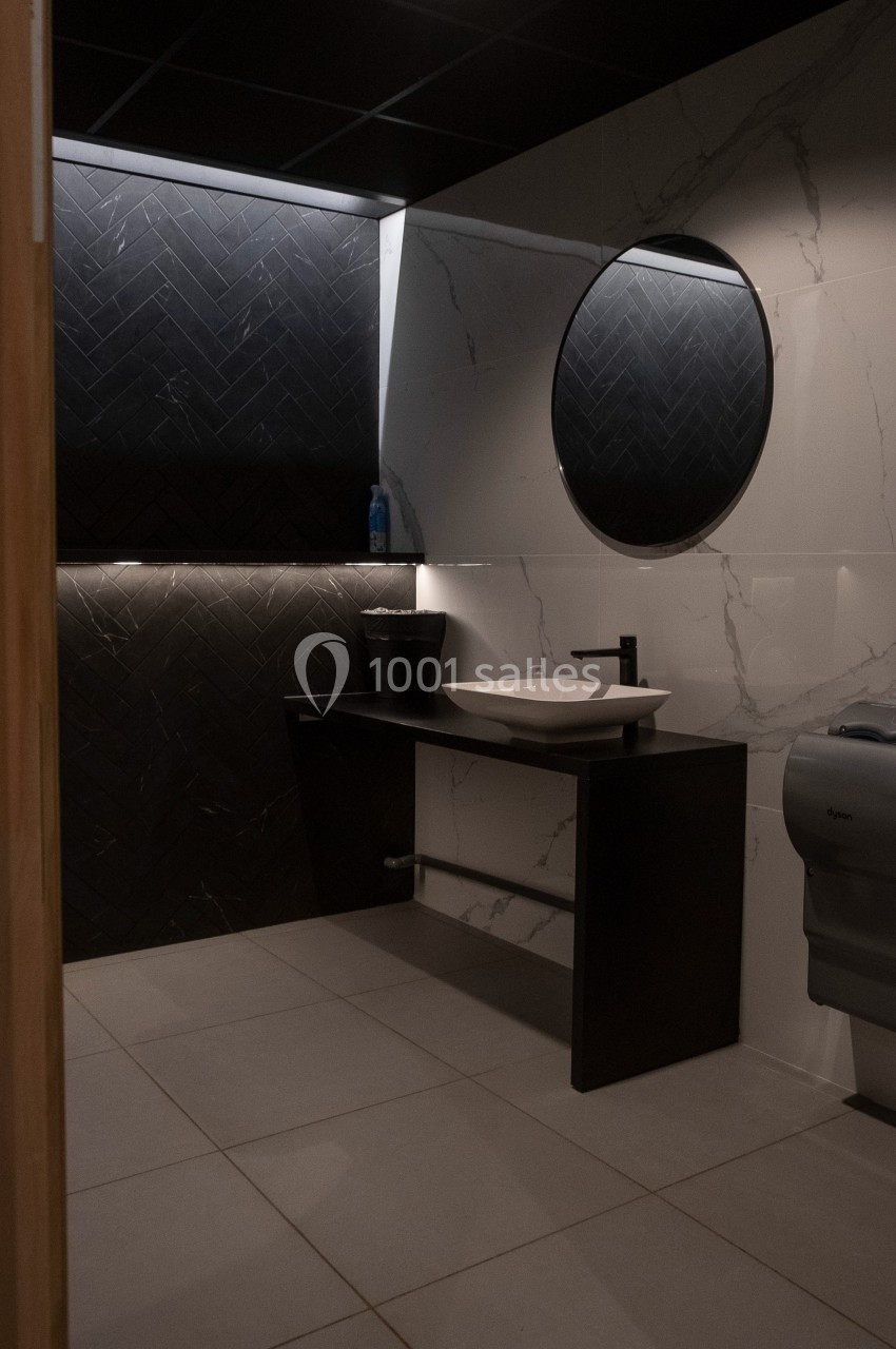 Salle de bain moderne avec lavabo, miroir rond, mur en carrelage noir et blanc, et éclairage tamisé.