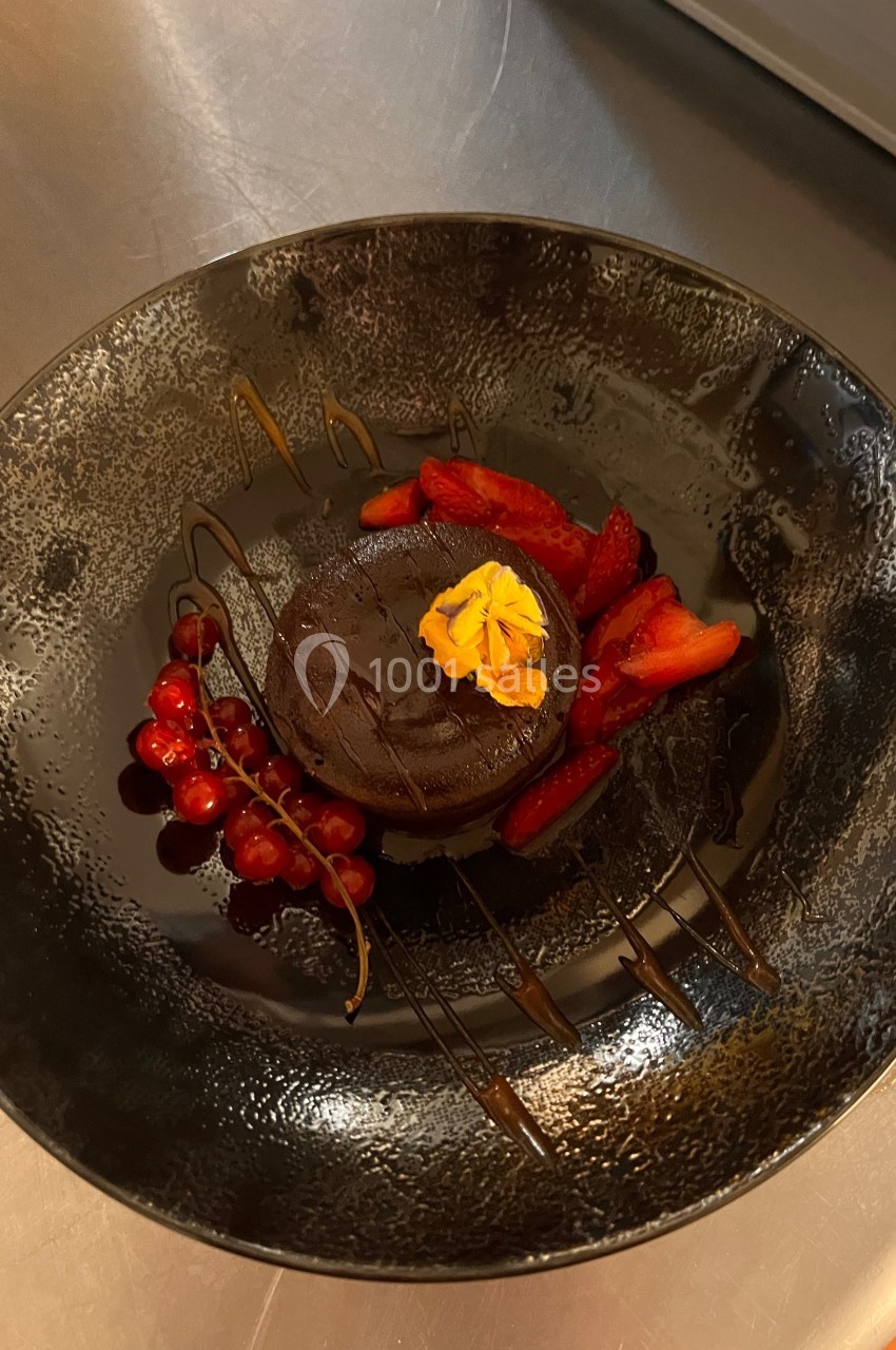 Dessert au chocolat décoré de fraises, groseilles et une fleur comestible, présenté sur une assiette noire.