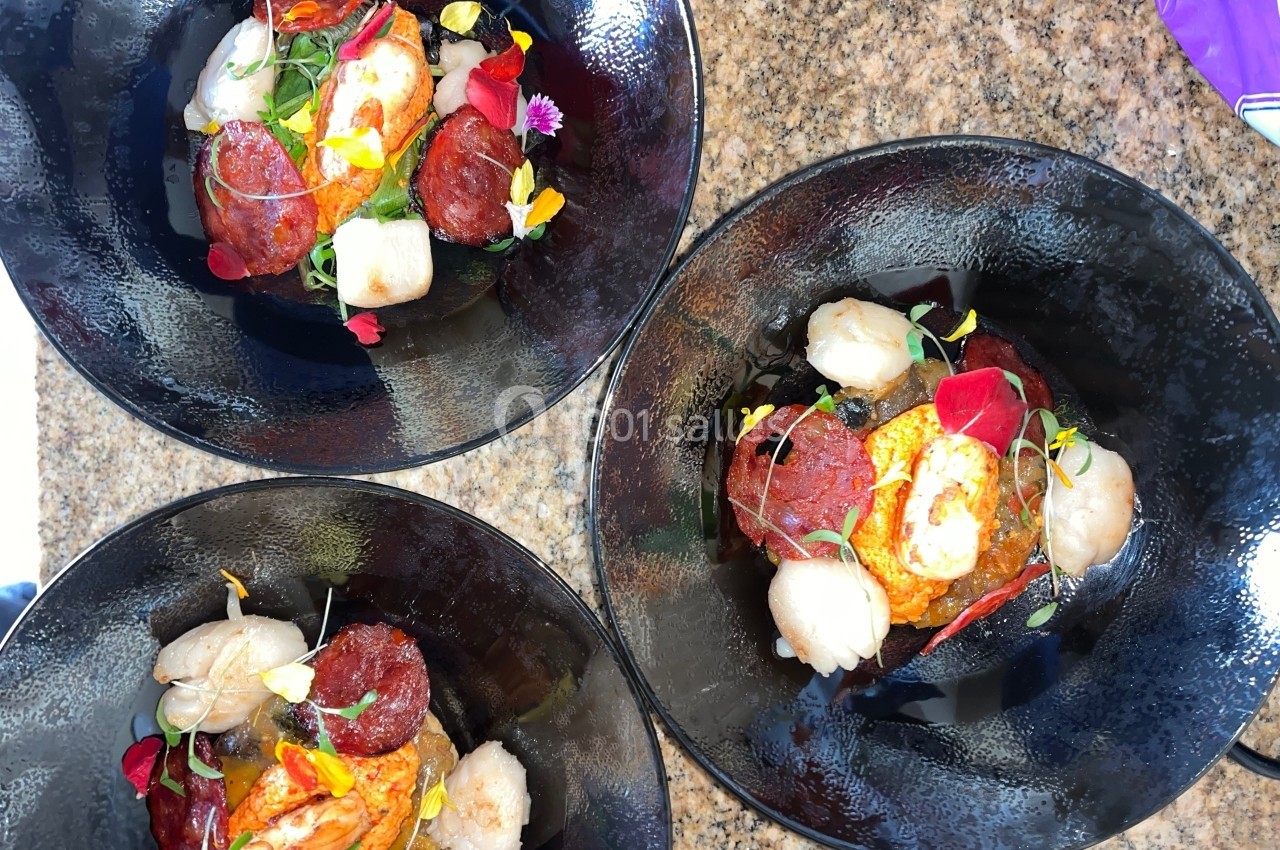 Trois assiettes noires contenant des pétoncles, des tranches de chorizo, des légumes et des fleurs comestibles.