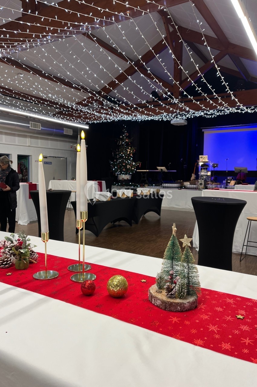 Salle décorée pour les fêtes avec guirlandes lumineuses, table rouge ornée de bougies et mini sapins, scène en arrière-plan.