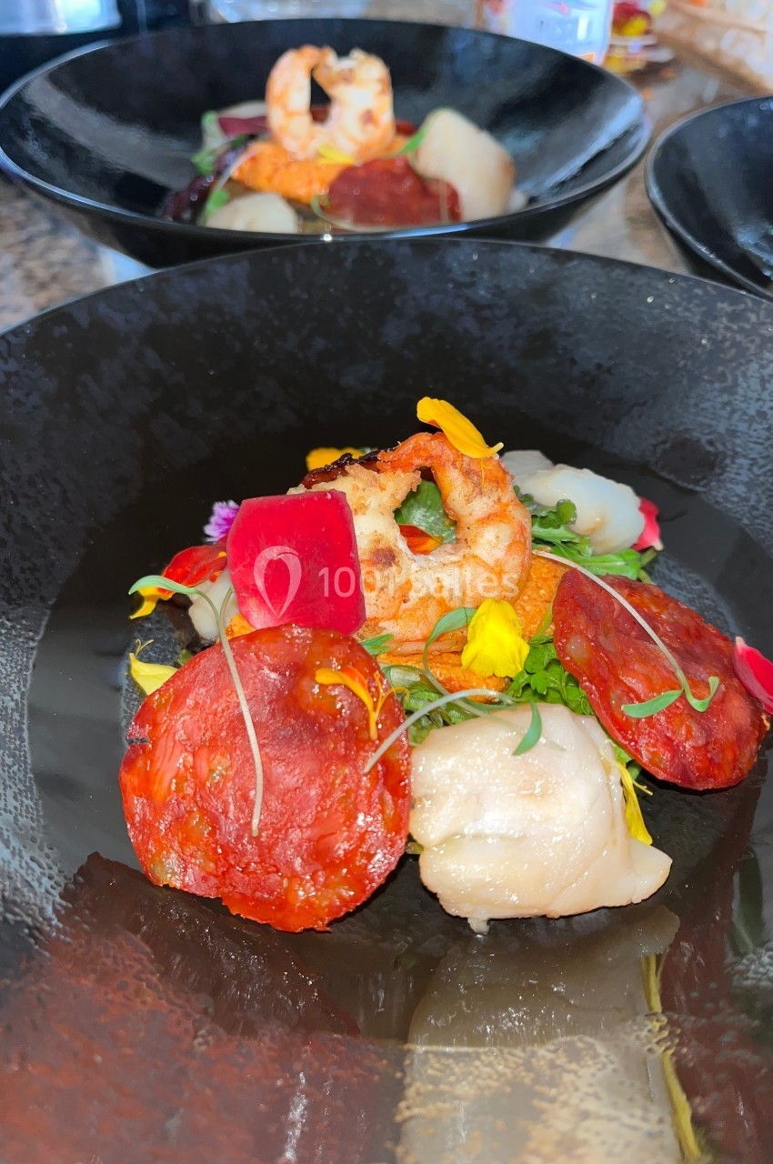 Assiette gastronomique composée de crevettes, chorizo, légumes colorés et herbes fraîches, servie dans un bol noir.