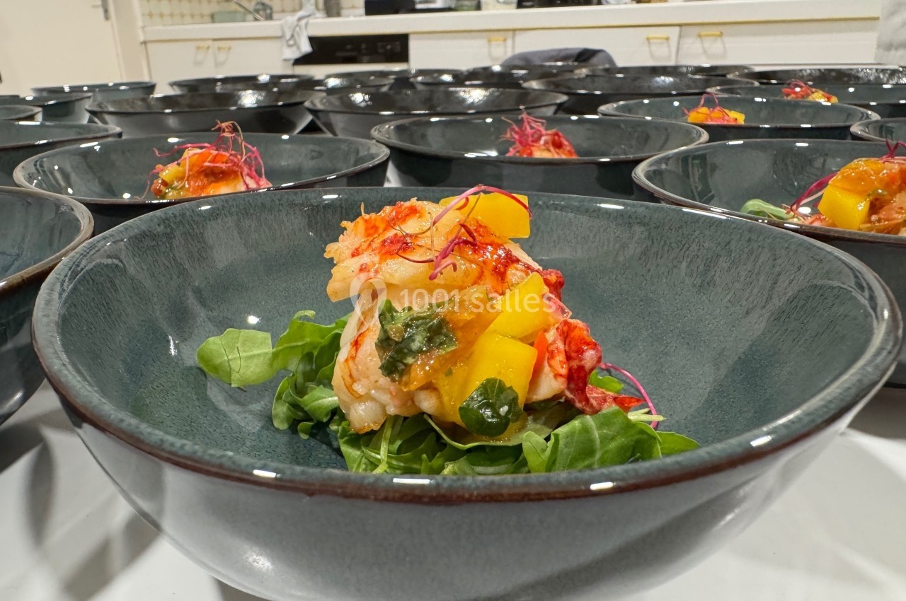 Plat dressé avec des morceaux de homard, dés de mangue et jeunes pousses sur un lit de salade verte, dans un bol foncé.