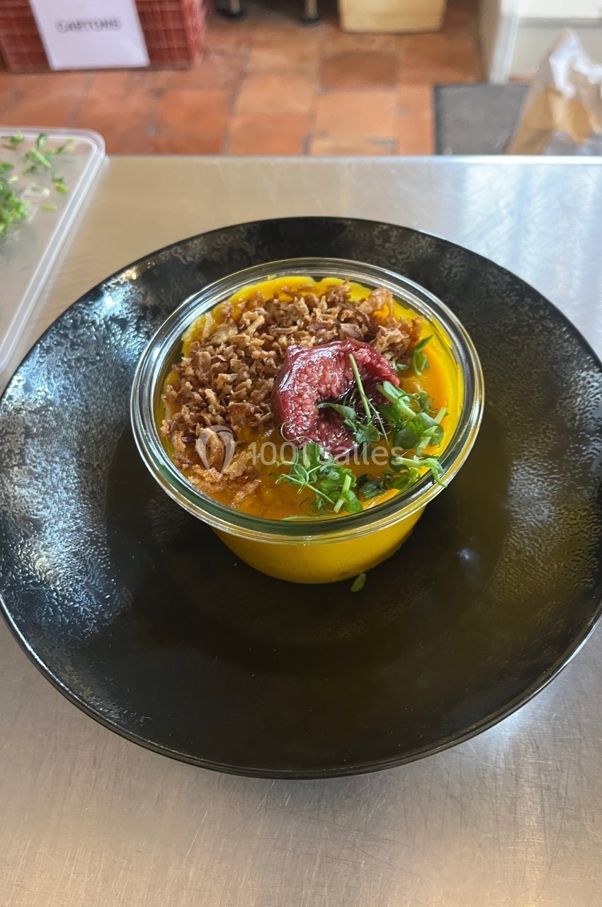 Bol de soupe orange garni de micro-pousses, oignons frits et morceaux de betterave, présenté sur une assiette noire.