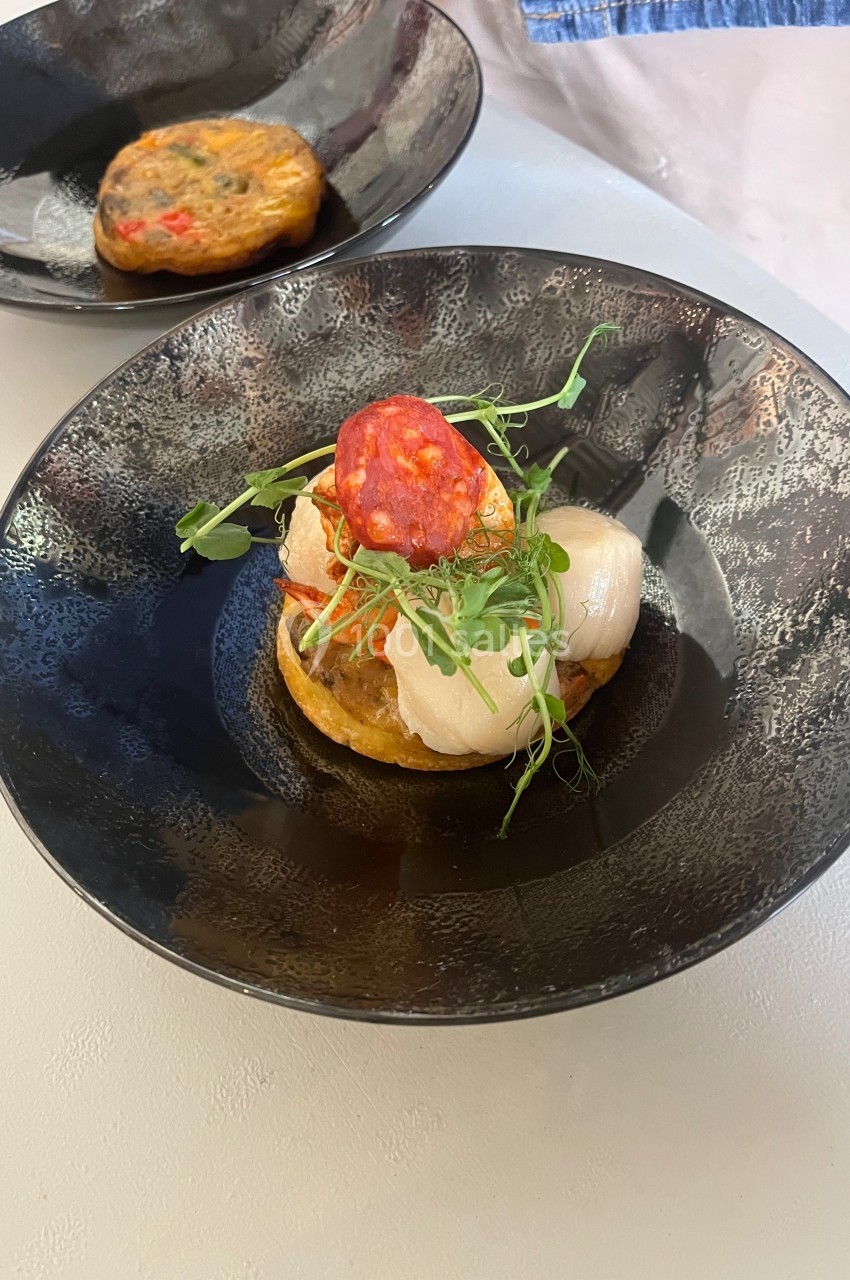 Plat dressé dans une assiette noire, composé de pétoncles, d'une galette de légumes, de verdure et d'une tranche de chorizo.