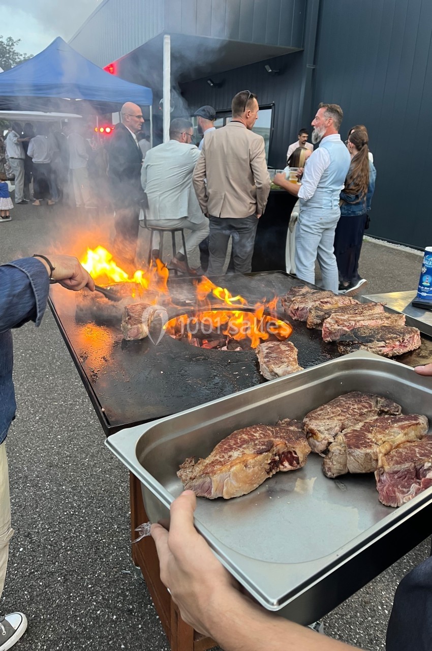 Personnes rassemblées autour d'un barbecue en plein air, avec des steaks en train de cuire sur une plancha enflammée.