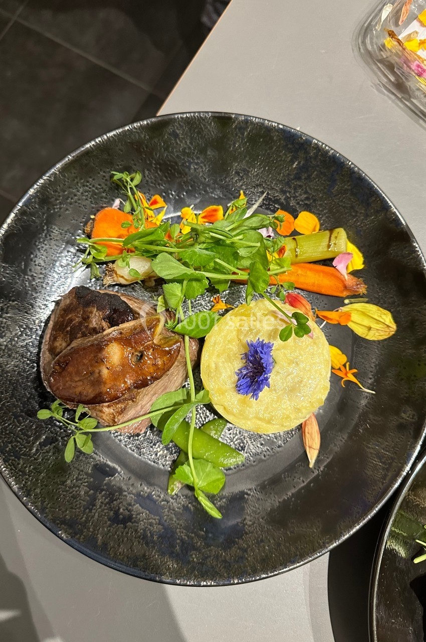 Assiette gastronomique avec viande, purée décorée d'une fleur bleue, légumes colorés et herbes fraîches.