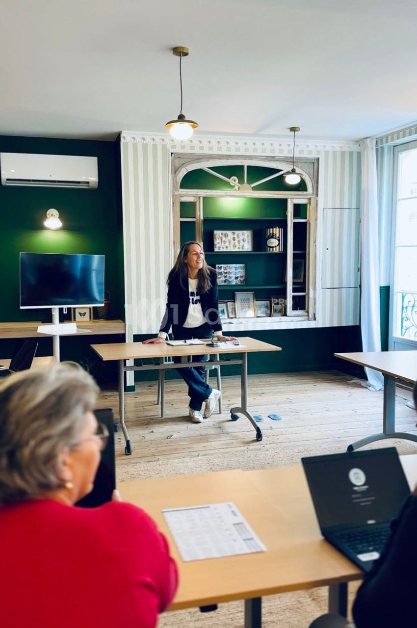 Une femme debout devant une table anime une présentation dans une salle lumineuse avec des participants assis.