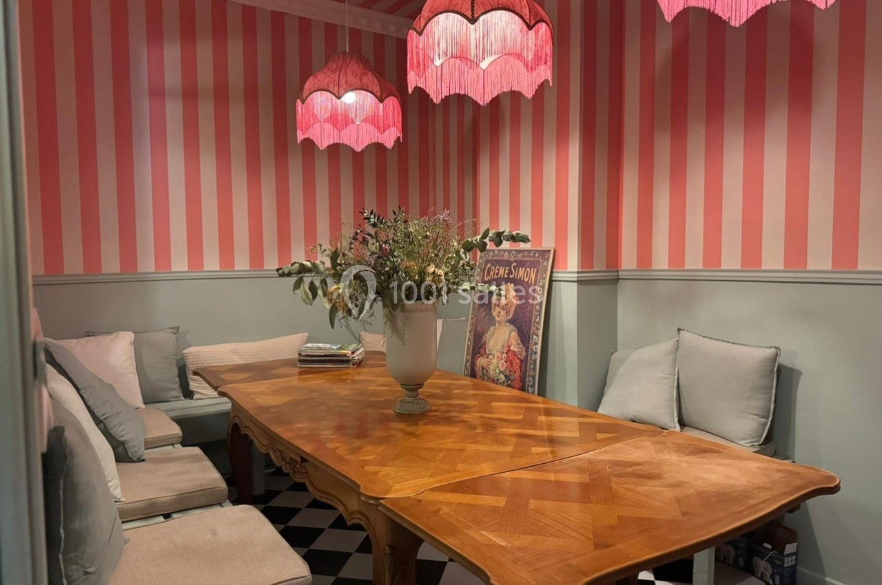 Salle à manger avec table en bois, banquettes et coussins, murs à rayures roses et blanches, luminaires roses suspendus.