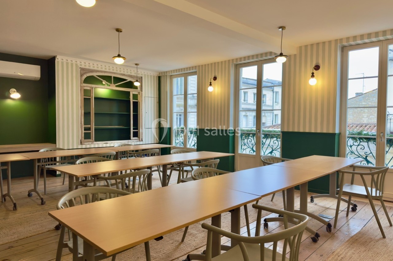 Salle lumineuse avec tables en bois, chaises modernes et grandes fenêtres donnant sur des bâtiments extérieurs.