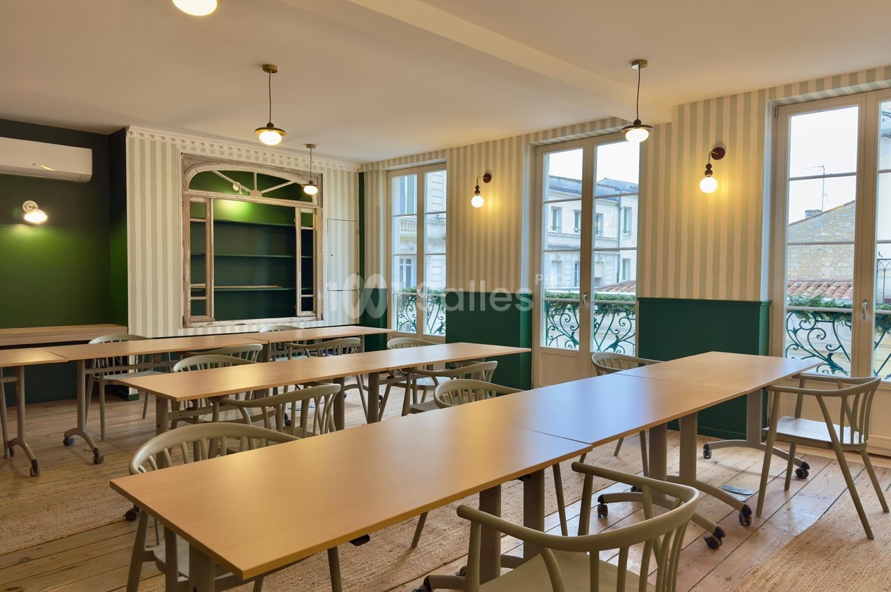 Salle lumineuse avec tables en bois, chaises modernes et grandes fenêtres donnant sur des bâtiments extérieurs.