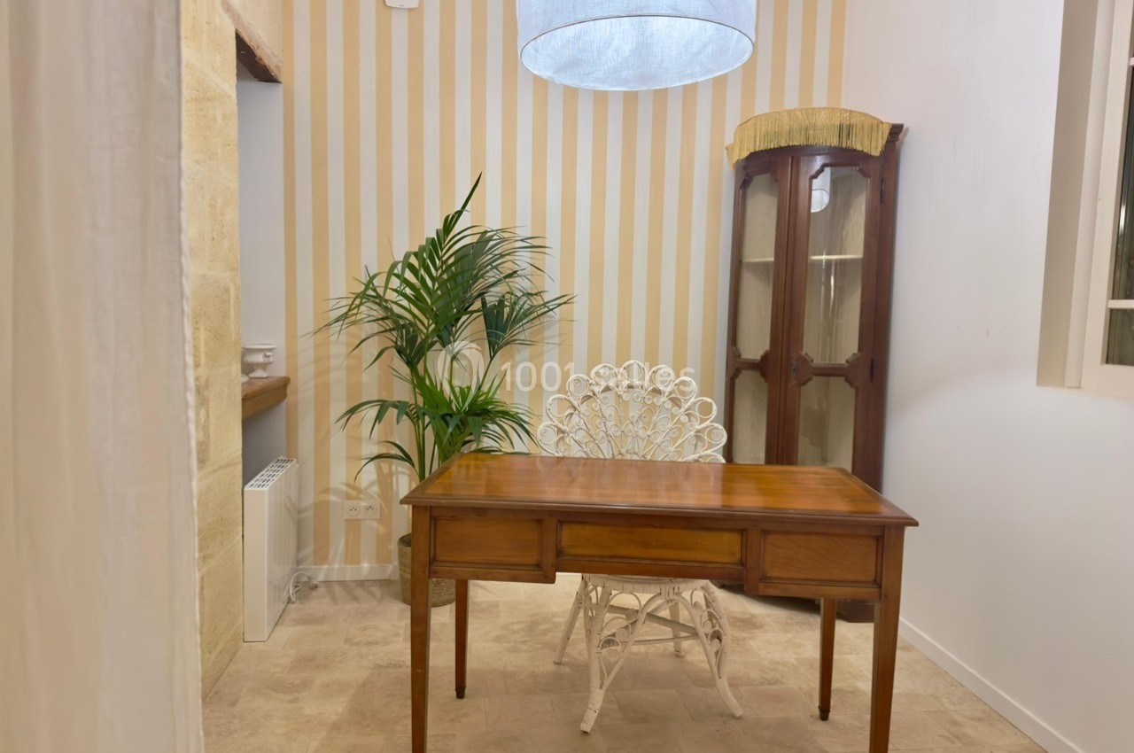 Bureau en bois avec chaise blanche en osier, placé dans une pièce lumineuse aux murs rayés et décorée d'une plante verte.
