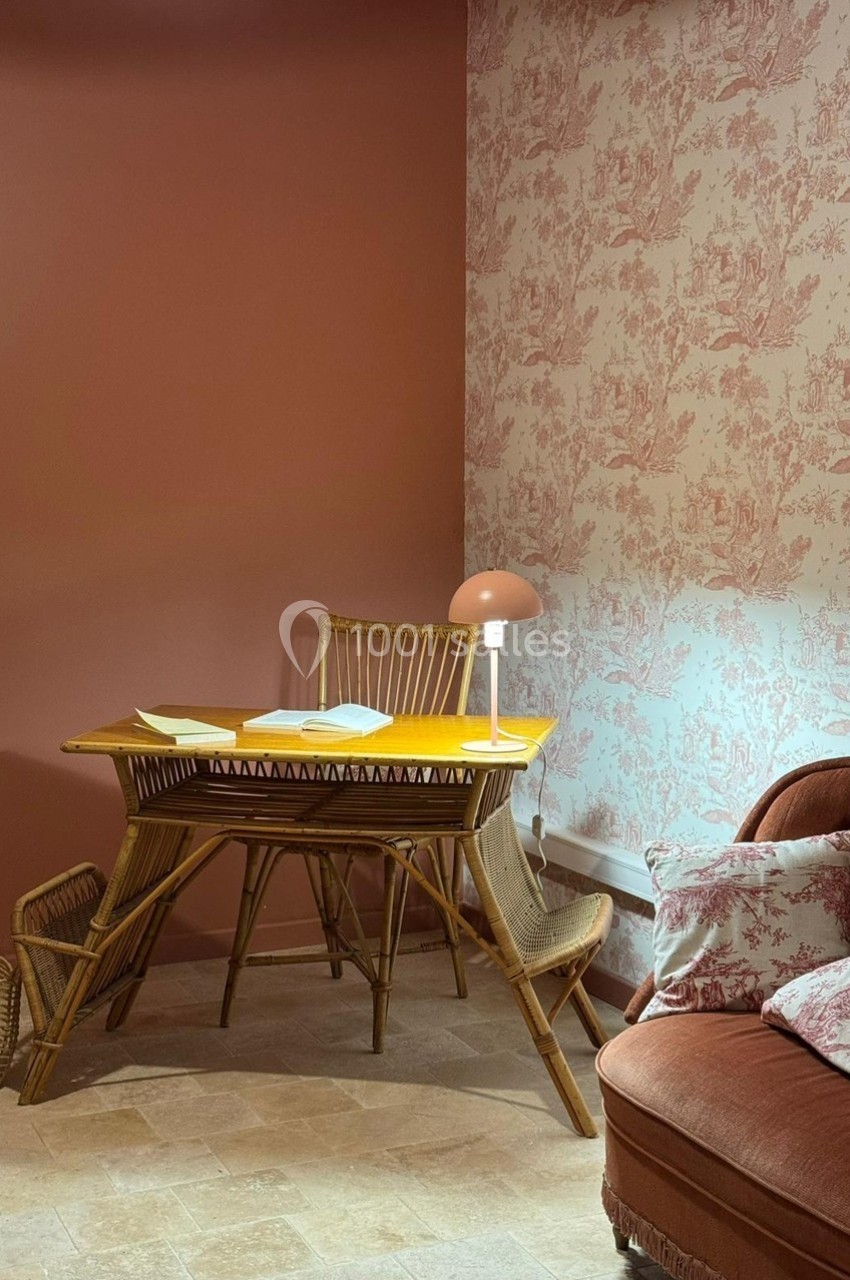 Coin bureau avec table en rotin, lampe allumée, mur rose uni et papier peint à motifs, ambiance chaleureuse.
