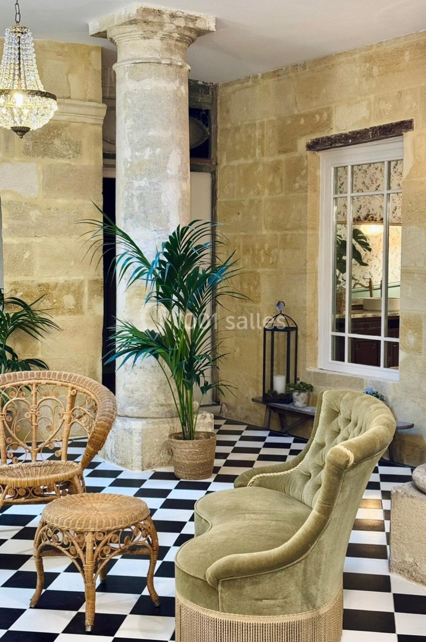 Espace intérieur avec sol en damier, fauteuil vert, meubles en rotin, plantes et colonnes en pierre.