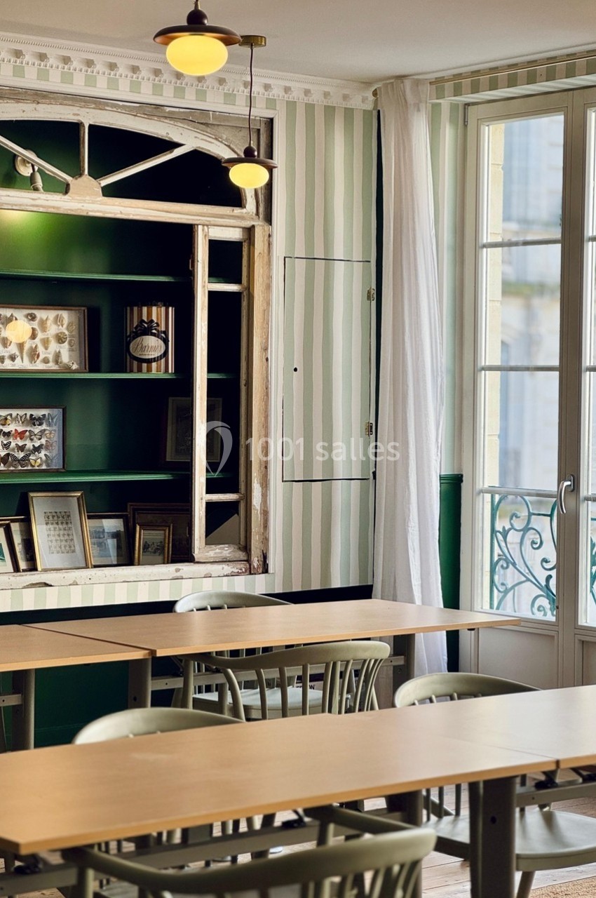 Salle lumineuse avec tables en bois, chaises vertes, étagères décorées et fenêtre donnant sur un balcon en fer forgé.