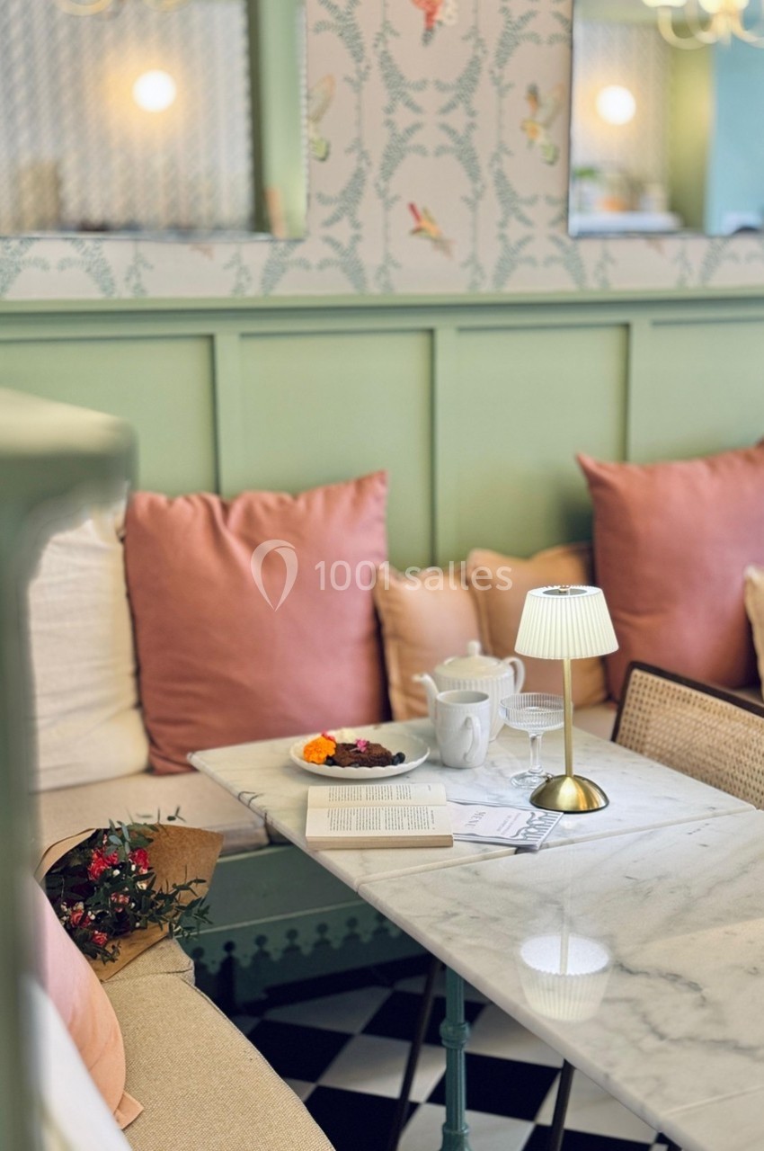 Coin salon cosy avec banquette pastel, coussins roses, table en marbre, lampe, bouquet de fleurs et tasse de thé.