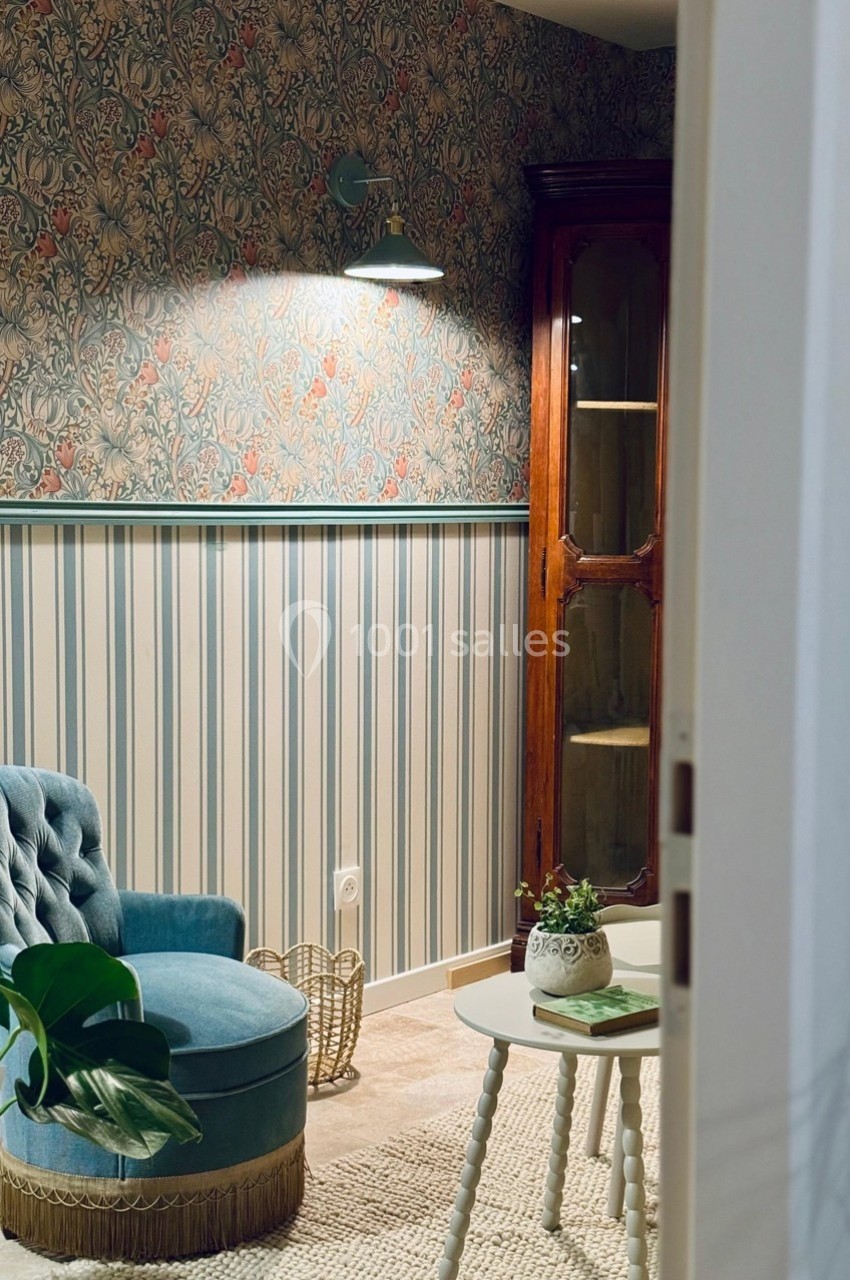 Fauteuil bleu et table basse dans un salon au style vintage avec papier peint fleuri et armoire en bois.