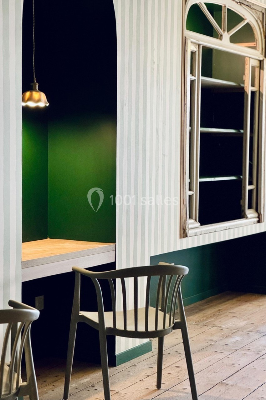 Espace intérieur avec murs verts et rayés, table en bois, chaise et étagère encadrée par une fenêtre ancienne.