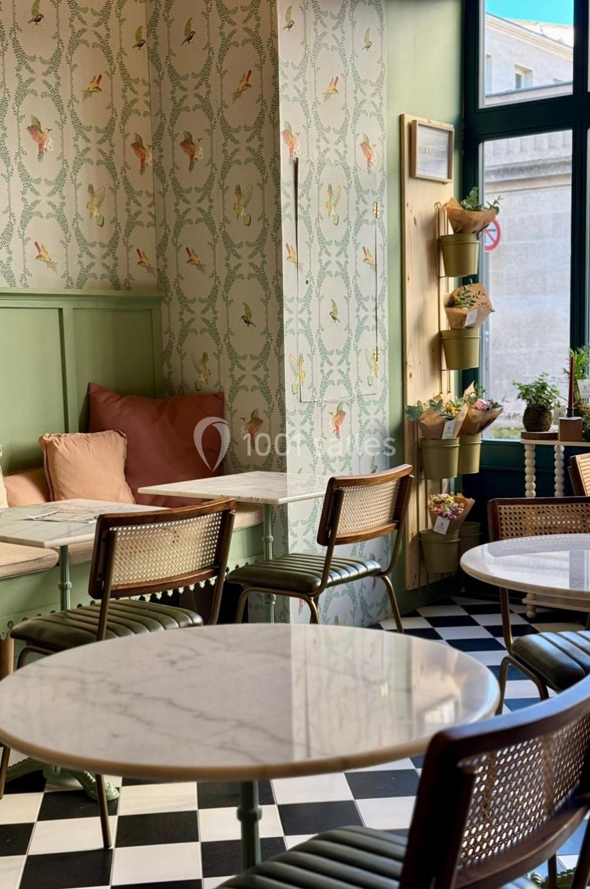 Salle de café lumineuse avec tables en marbre, chaises en rotin, papier peint floral et sol en damier noir et blanc.