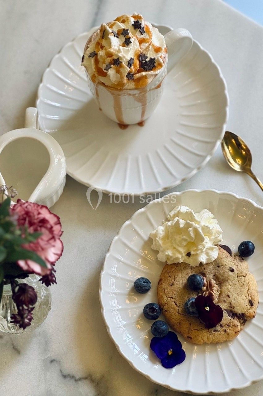 Tasse de chocolat chaud avec chantilly et caramel, accompagnée d'un cookie aux myrtilles et d'une fleur comestible.