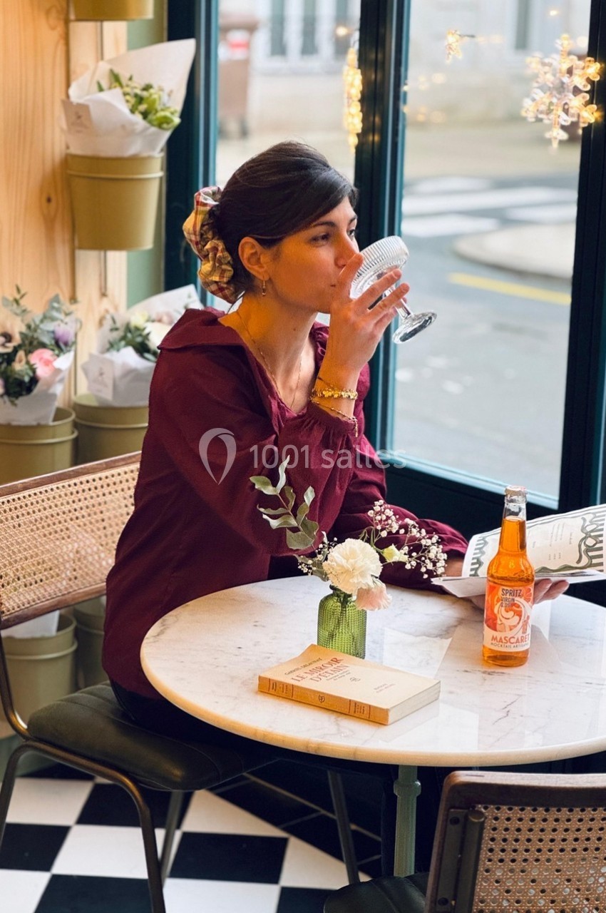 Une femme assise dans un café, buvant dans un verre, avec un livre et une bouteille posés sur une table.