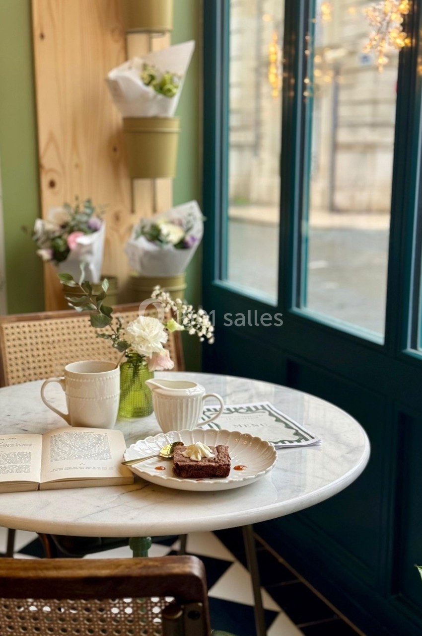Table ronde en marbre avec deux tasses, un dessert et un livre, près d'une fenêtre lumineuse et de fleurs décoratives.