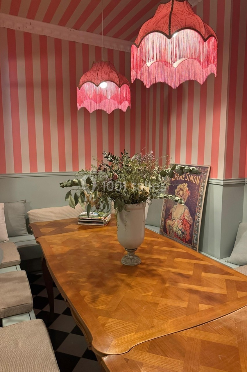 Salle à manger avec table en bois, banquettes rembourrées, lampes à franges roses et vase de fleurs au centre.