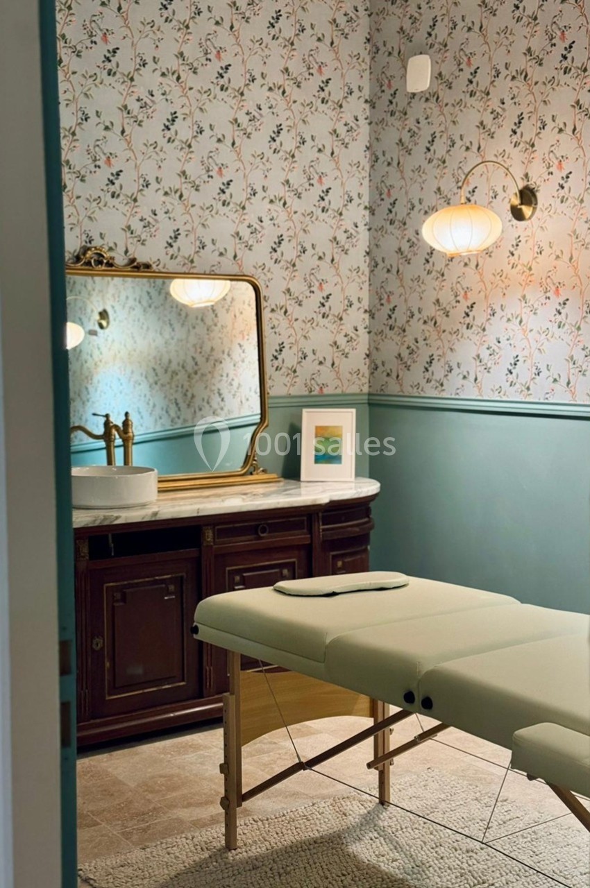 Salle de soins avec table de massage pliable, meuble ancien, miroir doré et mur à motifs floraux.
