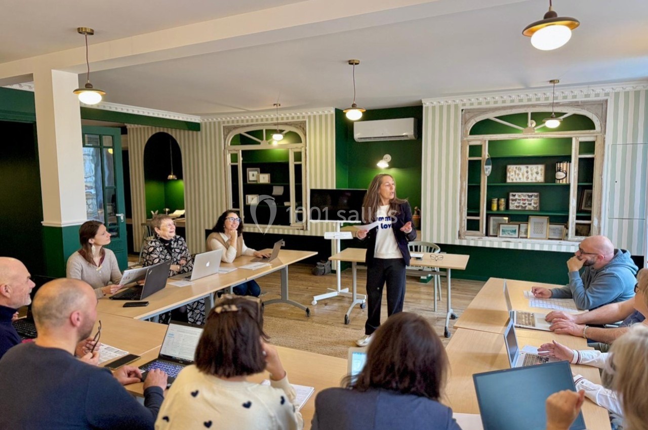 Une femme présente devant un groupe de personnes assises autour de tables avec des ordinateurs dans une salle lumineuse.