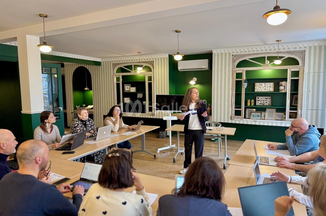 Une femme présente devant un groupe de personnes assises autour de tables avec des ordinateurs dans une salle lumineuse.