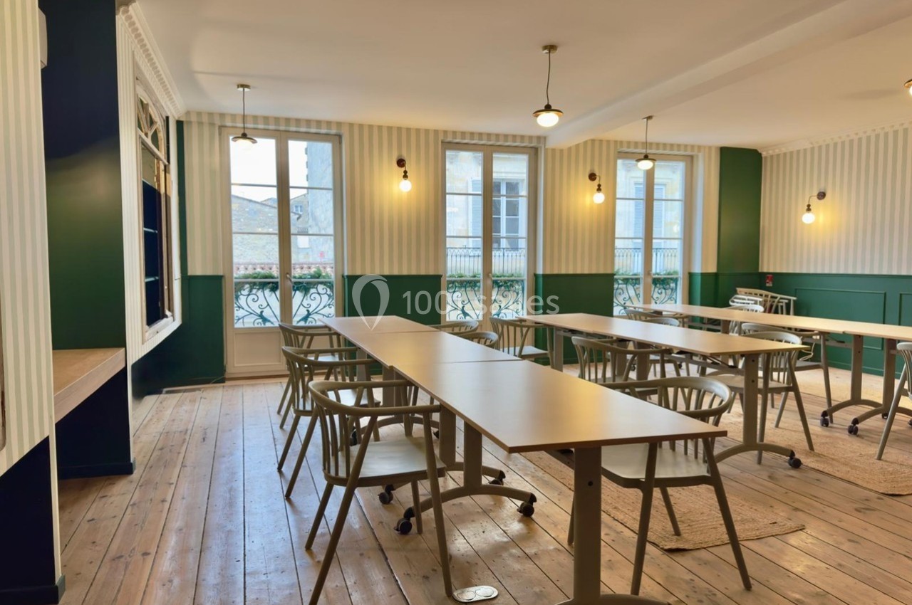 Salle lumineuse avec parquet en bois, tables et chaises alignées, murs verts et fenêtres donnant sur l'extérieur.