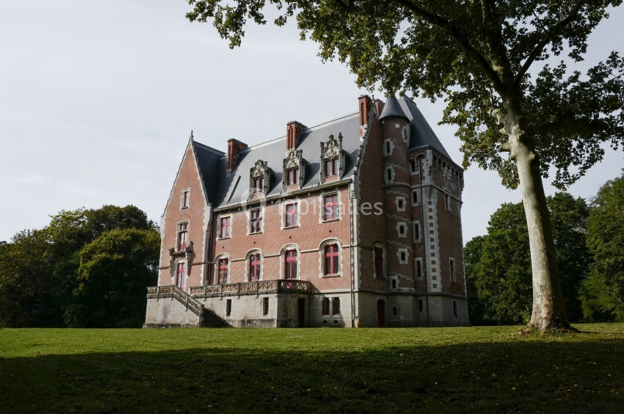 Château en briques rouges avec des fenêtres à meneaux, entouré d'un parc arboré sous un ciel clair.