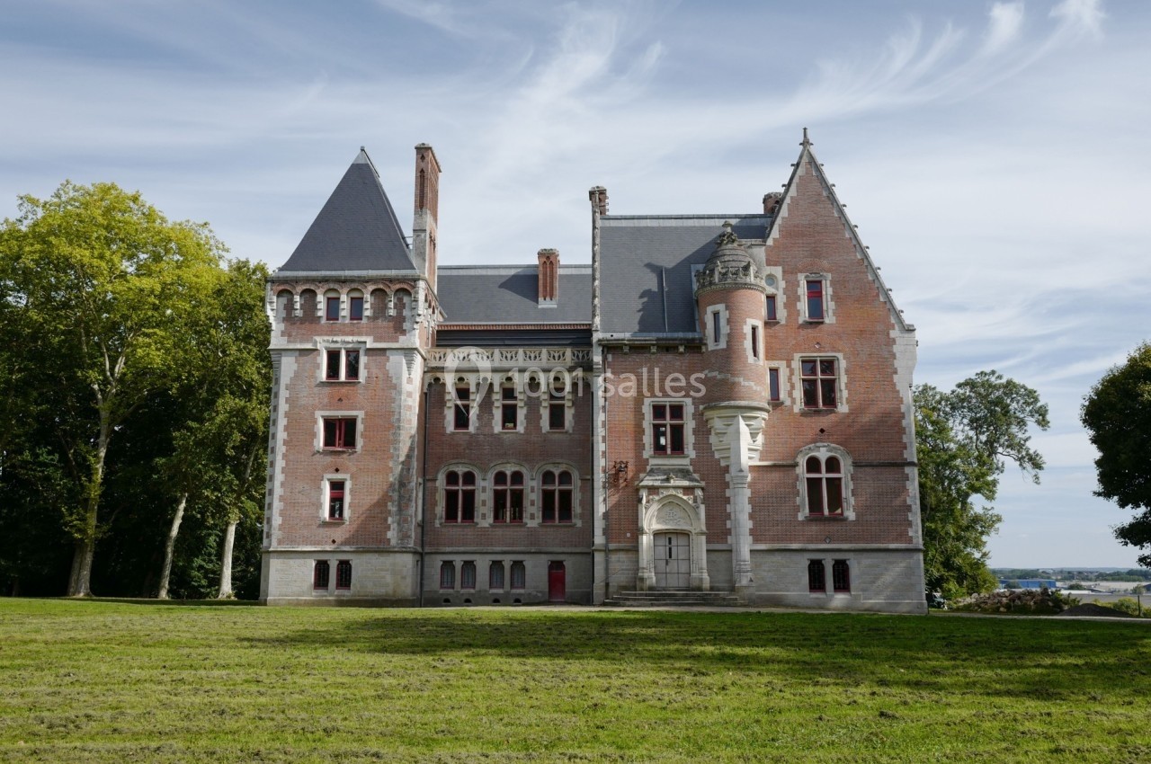 Façade d'un château en briques rouges avec des tours et des fenêtres gothiques, entouré d'arbres et d'une pelouse.