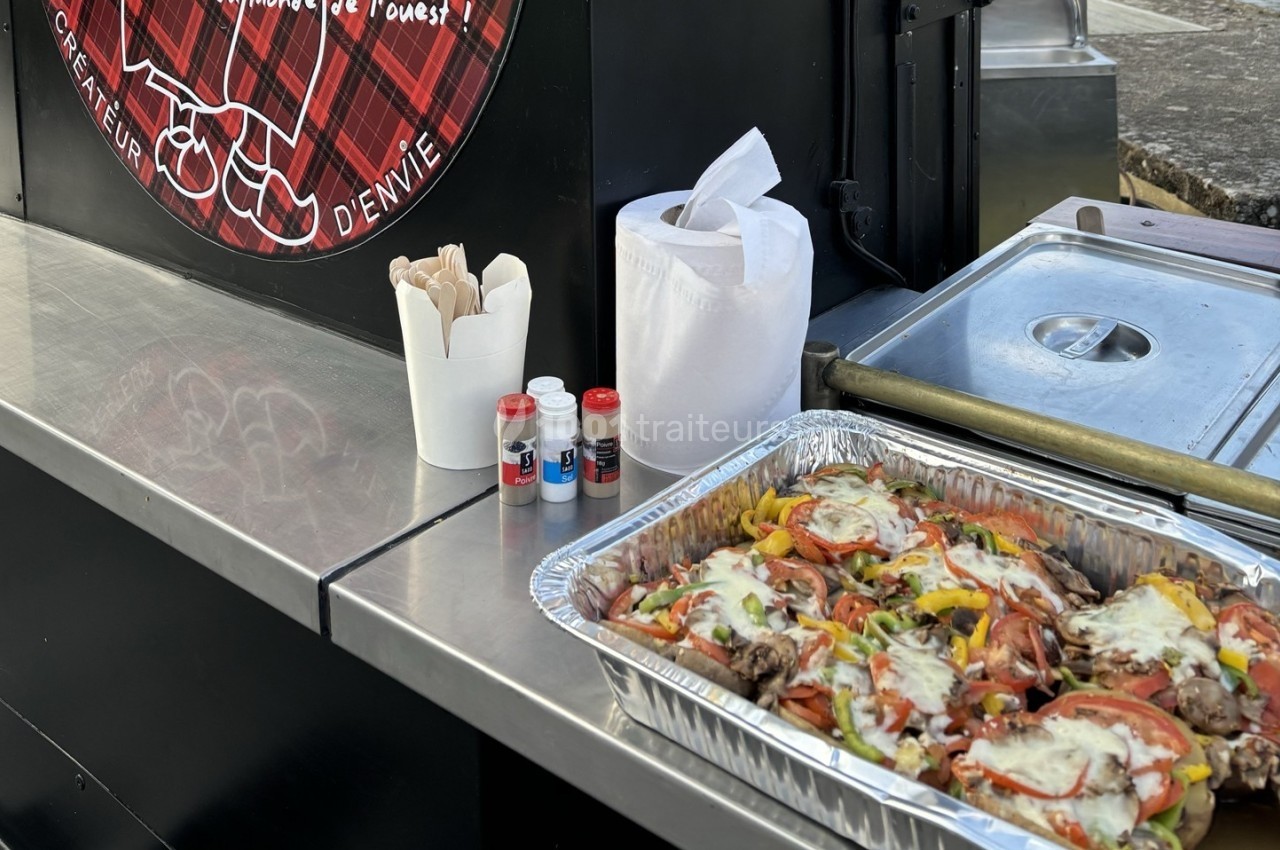 Plateau en aluminium contenant un plat de viande, légumes et fromage fondu, posé sur un stand de restauration mobile.