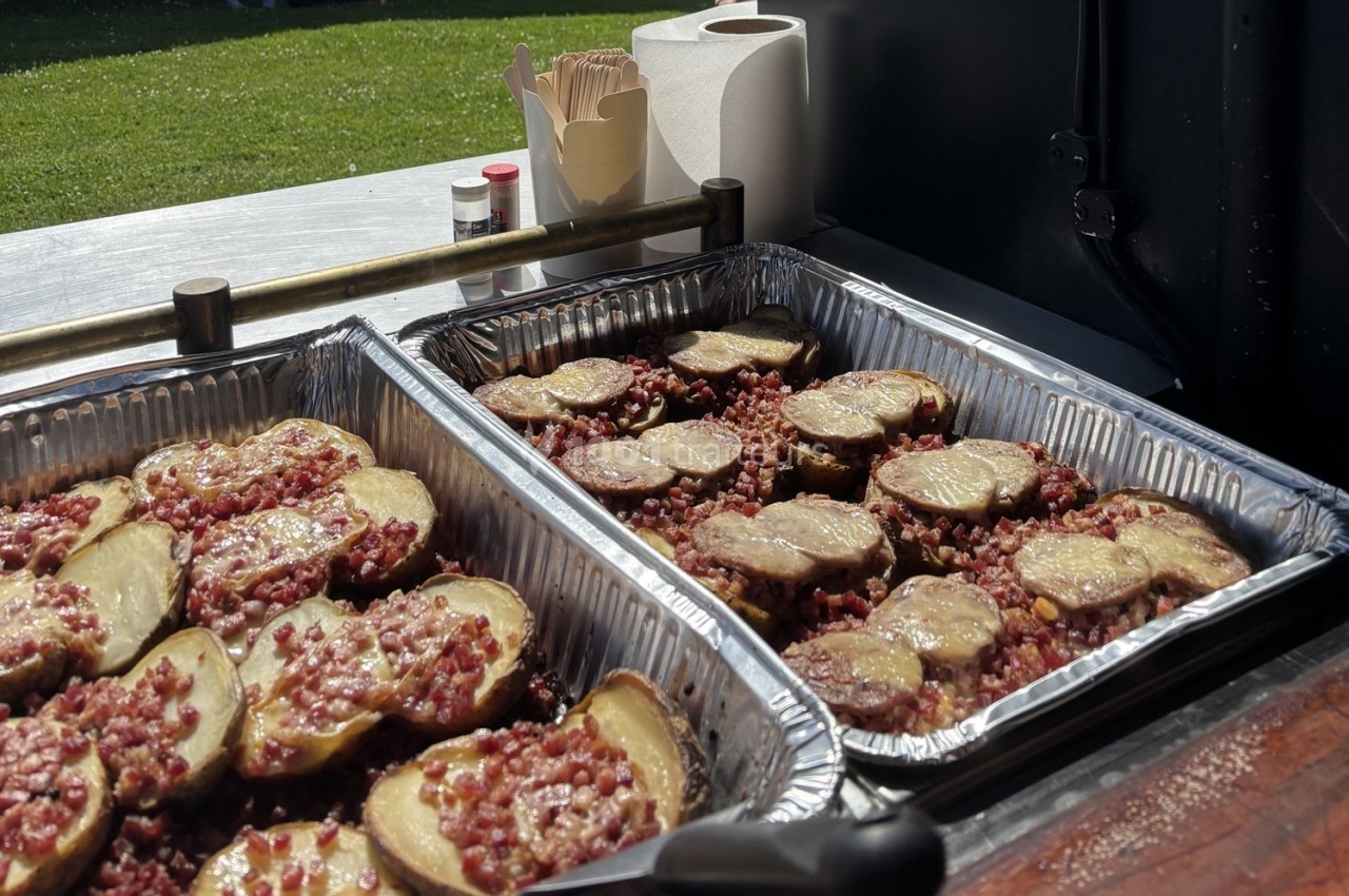 Pommes de terre garnies de fromage fondu et de lardons, présentées dans des plats en aluminium sur un stand en extérieur.