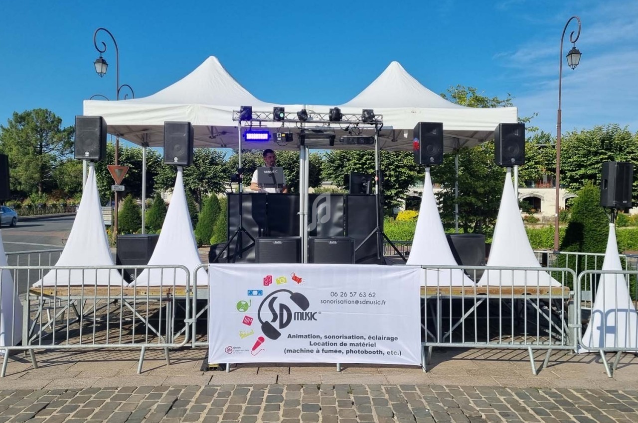 Scène extérieure avec un DJ sous une tente blanche, entourée de barrières et d'équipements d'animation.
