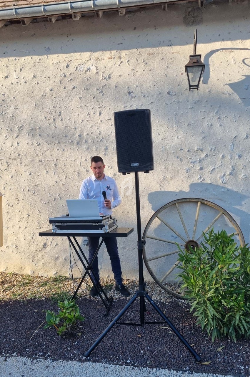 Un homme debout devant une table avec un ordinateur et un micro, près d'une enceinte, en extérieur.